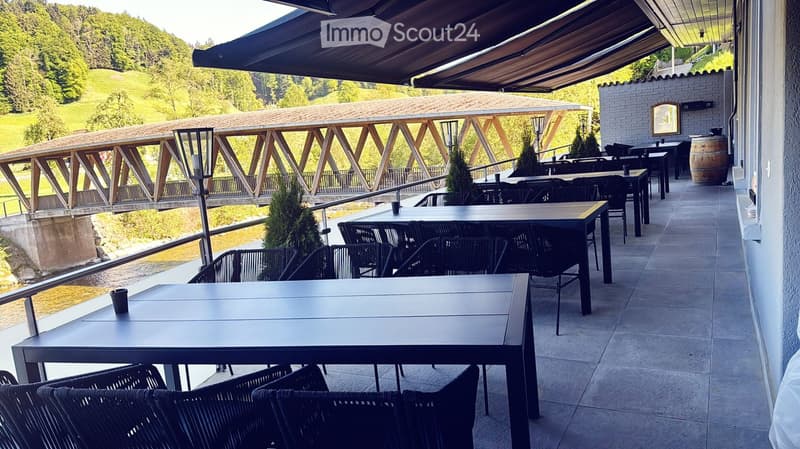 Outdoor terrace with tables and chairs, overlooking a river and wooden bridge, with a covered area and scenic view.
