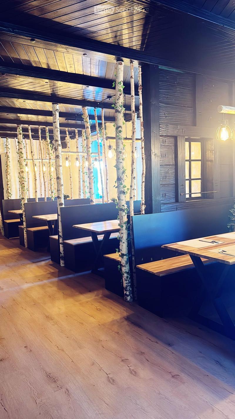 Restaurant interior with wooden tables, benches, and stools arranged around birch tree trunks, warm lighting, and wooden flooring.