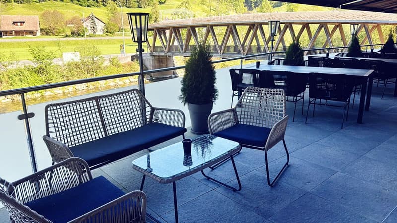 Outdoor terrace with wicker furniture, glass table, and scenic view of a river and bridge.