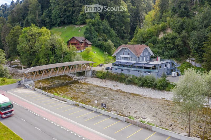 A large, modern building with a grey facade and red-tiled roof, situated near a river with a wooden bridge crossing it. The building has multiple windows and a balcony area, surrounded by lush green trees and a parking lot nearby.