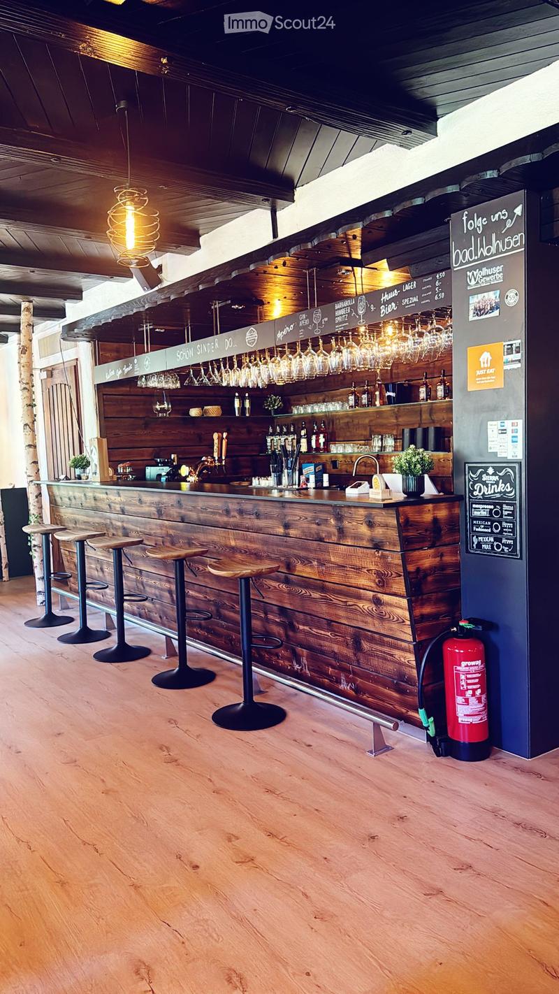 Wooden bar counter with hanging wine glasses, bottles, and stools, with a fire extinguisher nearby.