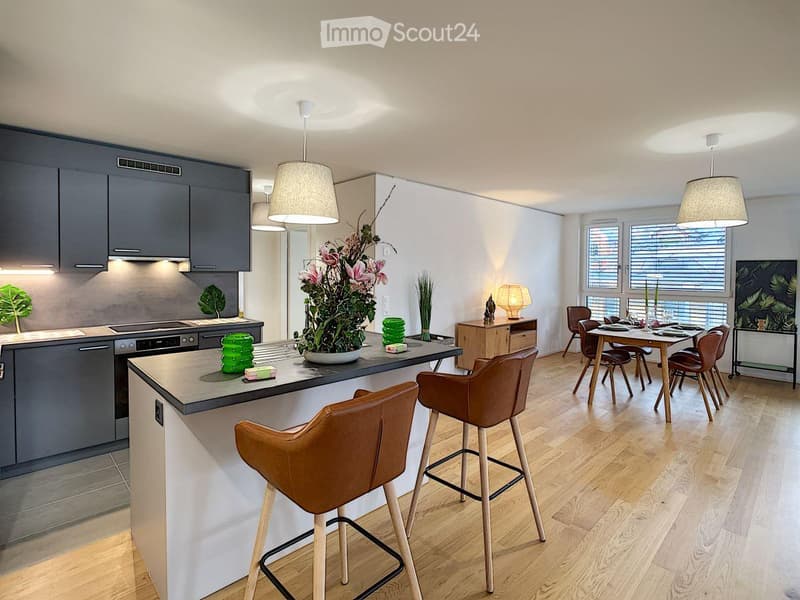 Modern kitchen with wooden floor, gray cabinets, and a large island with bar stools. The kitchen includes a stove, oven, and a sink. There is a dining area with a table and chairs. A plant and flowers are on the island, and a lamp is on the side table.