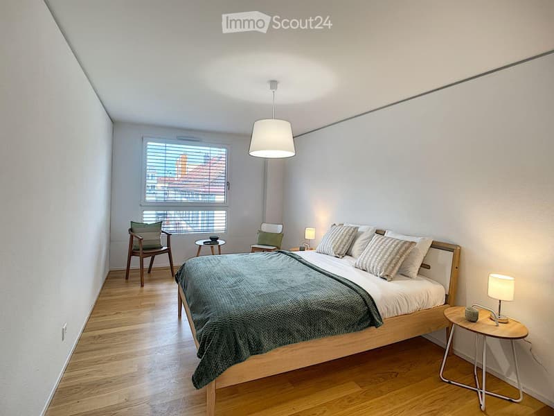 Spacious bedroom with wooden floor, white walls, and a large bed. Two nightstands with lamps and a chair near the window. A pendant lamp above the bed.