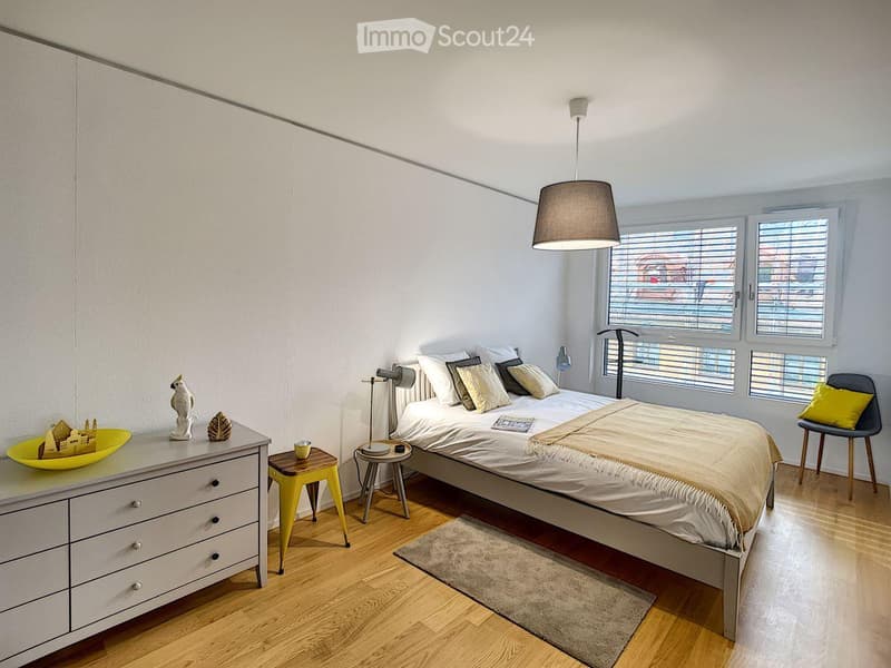 A bedroom with a bed, nightstands, a chair, and a dresser. The bed has a white sheet and a beige blanket. The nightstands have lamps and magazines. The dresser has a yellow bowl and a figurine. The room has a wooden floor and white walls. The windows have