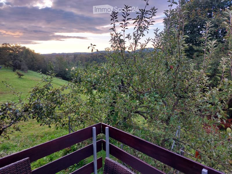 A balcony with a wooden railing and two wicker chairs, overlooking a grassy field with apple trees and a distant hill under a cloudy sky.