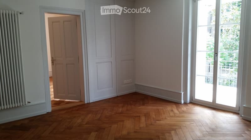 An empty room with white walls, wooden herringbone flooring, and two sets of glass doors. One set of doors leads to another room, and the other set opens to a balcony with a view of trees.