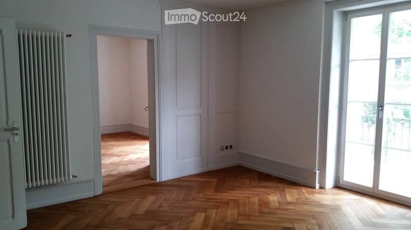 Empty room with white walls, wooden parquet flooring, and large windows. There is a radiator on the left wall and a door leading to another room.