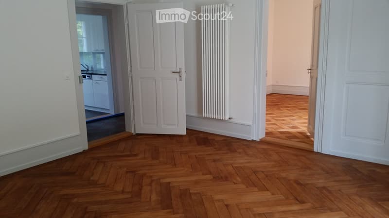 Empty room with wooden parquet flooring, white walls, and open doors leading to a kitchen and another room.