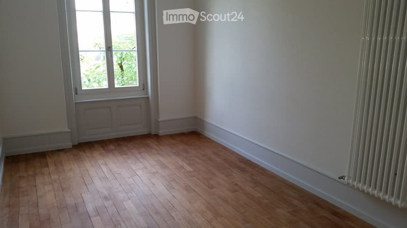 Completely empty room with wooden floors, white walls, and a window with a view of greenery outside.