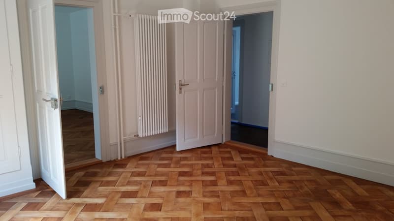 Empty room with wooden parquet flooring, white walls, and three open doors leading to other rooms. A radiator is mounted on the wall between two of the doors.