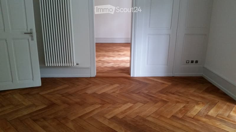 Empty room with wooden herringbone flooring, white walls, and two closed doors. A radiator is mounted on the wall on the left side.