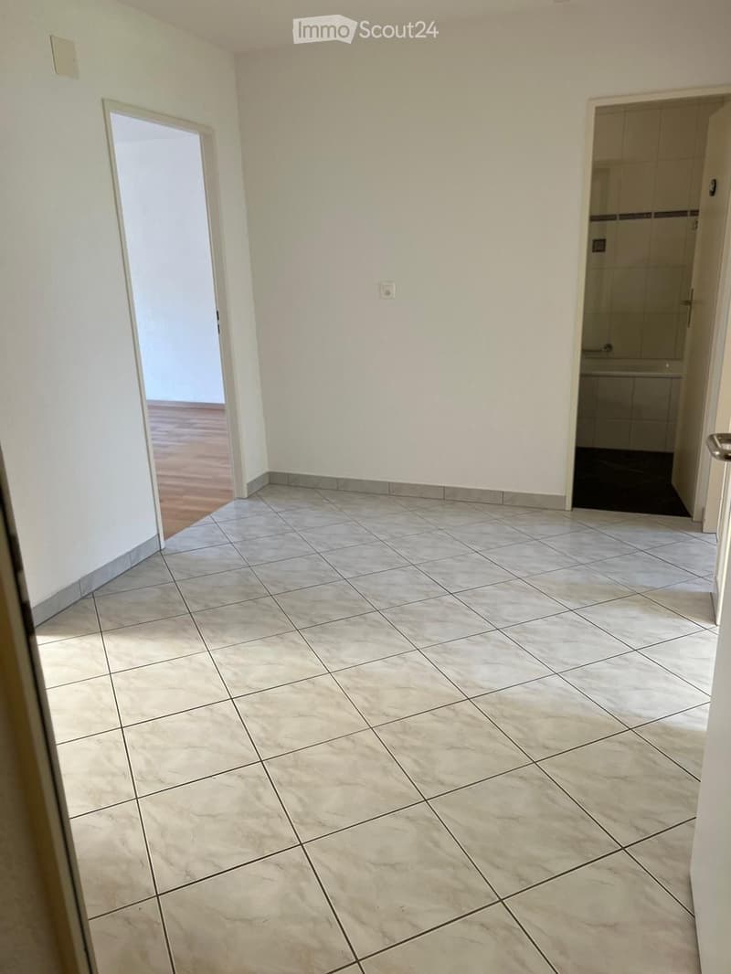 Empty room with white walls, white tiled floor, wooden floor in the background, two doors, one leading to a bathroom with bathtub