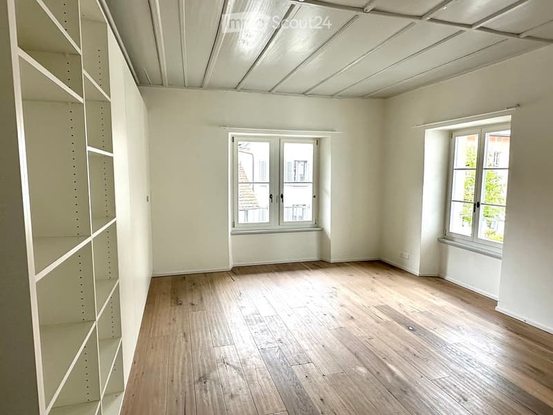 Empty room with wooden floors, white walls, and built-in shelves. Two windows with views of the outside.