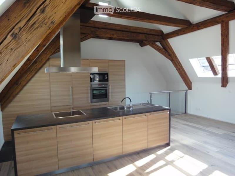 Modern attic kitchen with wooden beams, wooden cabinets, granite countertops, stainless steel sink, faucet, oven, and microwave.