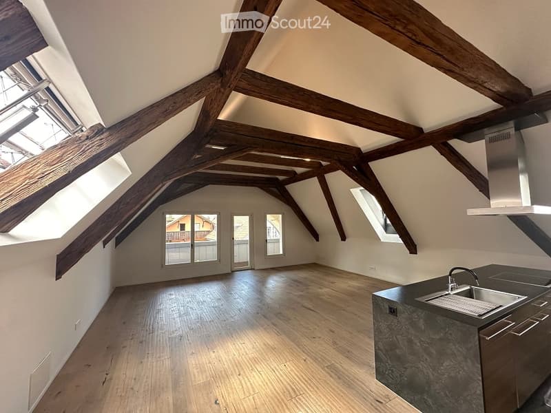 A spacious attic room with wooden beams on the ceiling and wooden floor, a kitchen area with a sink and cabinets, windows providing views, and white walls.