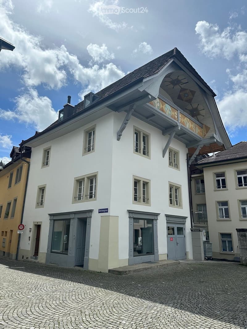 The building has 3 floors. The lower floor has large arched windows, the middle floor has smaller windows, and the top floor has slanted windows. The exterior of the building is white with brown and tan detailing. There are double doors at the entrance.