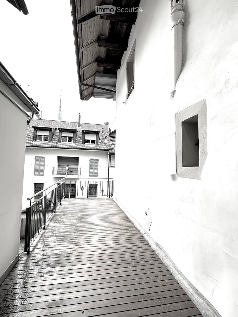 A black and white photograph of a building's exterior with a balcony. The building has a wooden walkway, a balcony with railings, and windows with shutters.