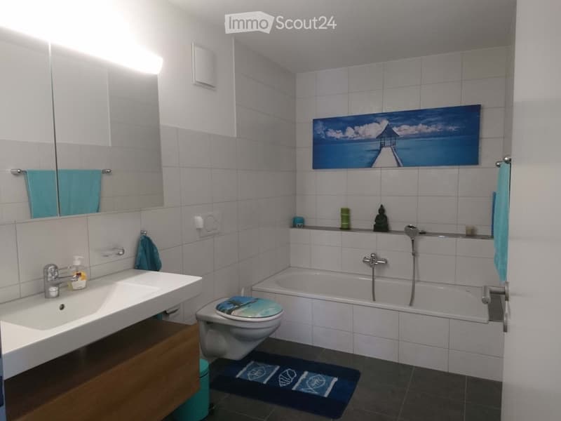 modern white tiles, white bathtub, white sink, mirror, painting, blue towels, green bottle, wooden cabinet