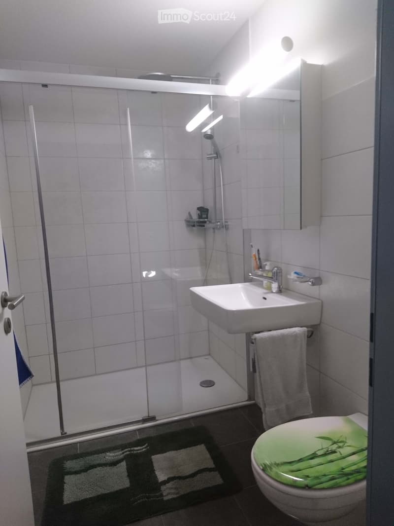 A white tiled bathroom with a glass walk-in shower, sink, and toilet. White tiles and white countertops, silver hardware.