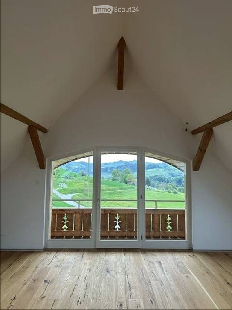 Empty room with wooden floor, white walls, and large glass doors with wooden frames. View of a green field and mountains.