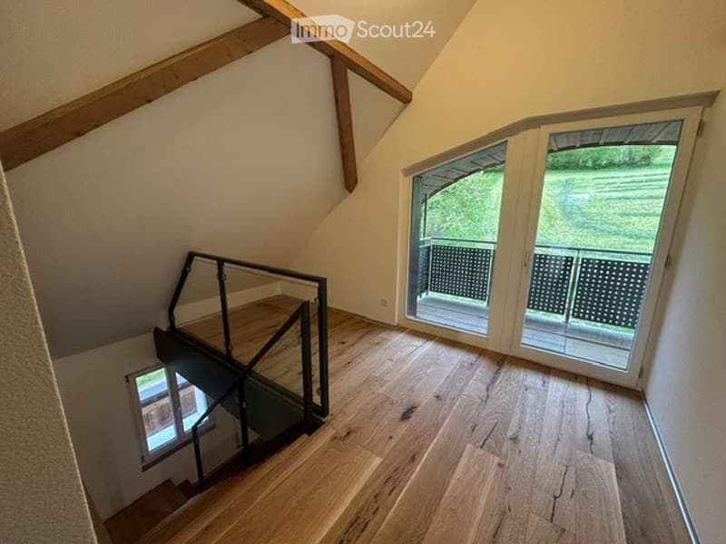 An empty attic room with wooden flooring, a staircase, and a balcony with a metal railing. The room has a glass door leading to the balcony and a small window on the left side. The walls are painted in a light color, and the ceiling has wooden beams. The