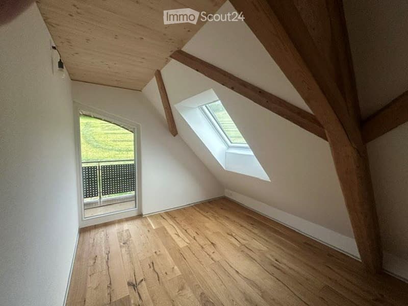 An empty attic room with wooden floors, white walls, wooden beams, a glass window, and a balcony with a metal railing.