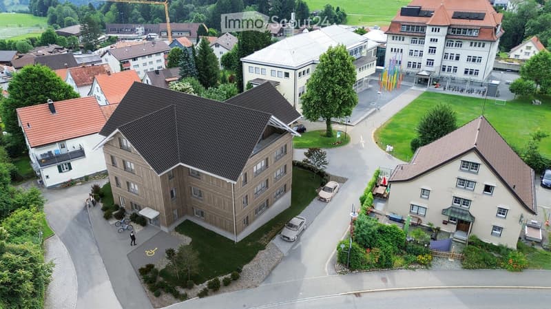 Aerial view of a large apartment building with multiple cars parked in front, surrounded by several houses and greenery.