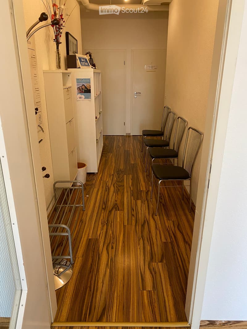 Other. {"visible_features":[],""type":"Corridor / Hallway",""objects":["chairs","cabinet","trash bin","vase","flowers","poster","picture frame","book","sign"],""description":"A hallway with wooden floors, a row of chairs, a cabinet with a vase of flowers,