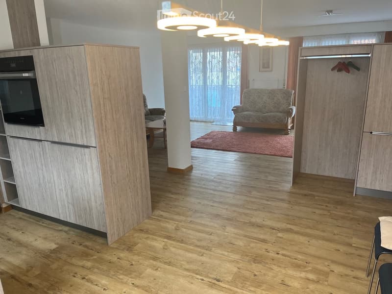 Furnished Room. A modern, open-concept living space with wooden flooring, a patterned sofa, a small table, and a built-in cabinet with an oven. The area is well-lit with large windows and hanging lights.