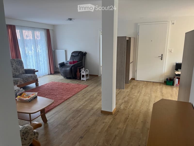 Furnished Room. Living room with wooden flooring, a red rug, a sofa, a coffee table, a recliner chair, and a small table with a laptop. There are large windows with curtains and a radiator near the window.