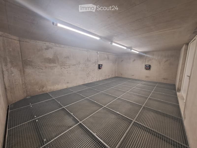 Empty concrete-walled room with a metal grated floor and fluorescent lighting, showing no furniture or equipment.