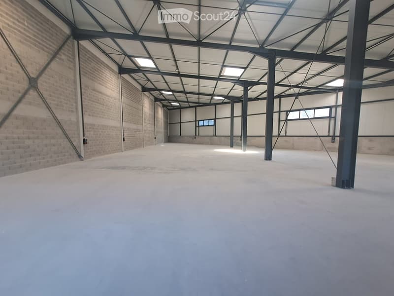 Empty industrial-style room with exposed brick walls, metal beams, and a concrete floor. The space is well-lit with natural light from large windows and a skylight.