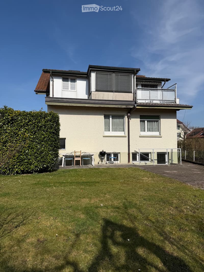 House. Two-story house with a brown roof, white walls, and a balcony on the second floor. The house has a fenced garden in front with a table and chairs.