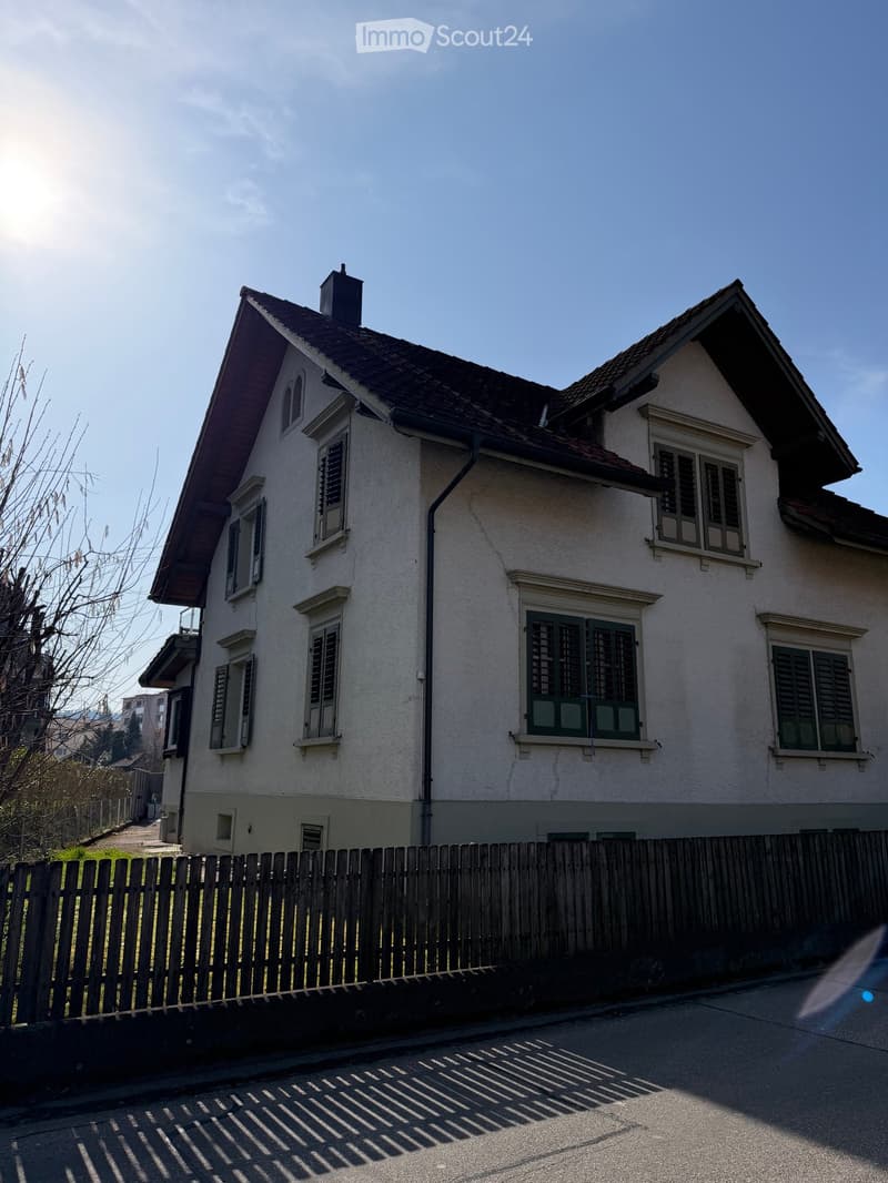 House. Two-story house with a brown roof, white walls, and green shutters on the windows. The house is surrounded by a wooden fence.
