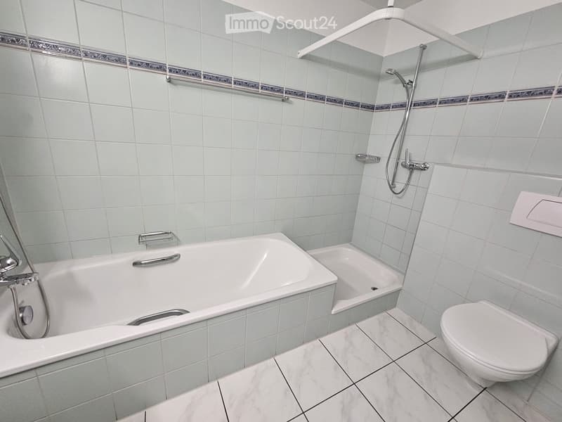 Modern bathroom with a large bathtub, separate shower area, and a toilet. Light blue tiles with decorative borders and marble flooring.
