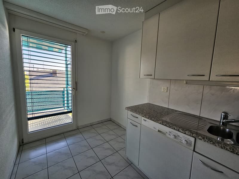 Modern kitchen with white cabinets, granite countertop, sink, and dishwasher. Features a large window with blinds leading to a balcony.