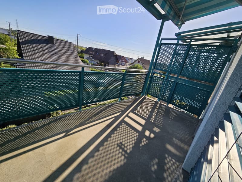 Balcony with green metal railing and patterned shadows from the railing on the concrete floor, overlooking a residential area with houses and trees.