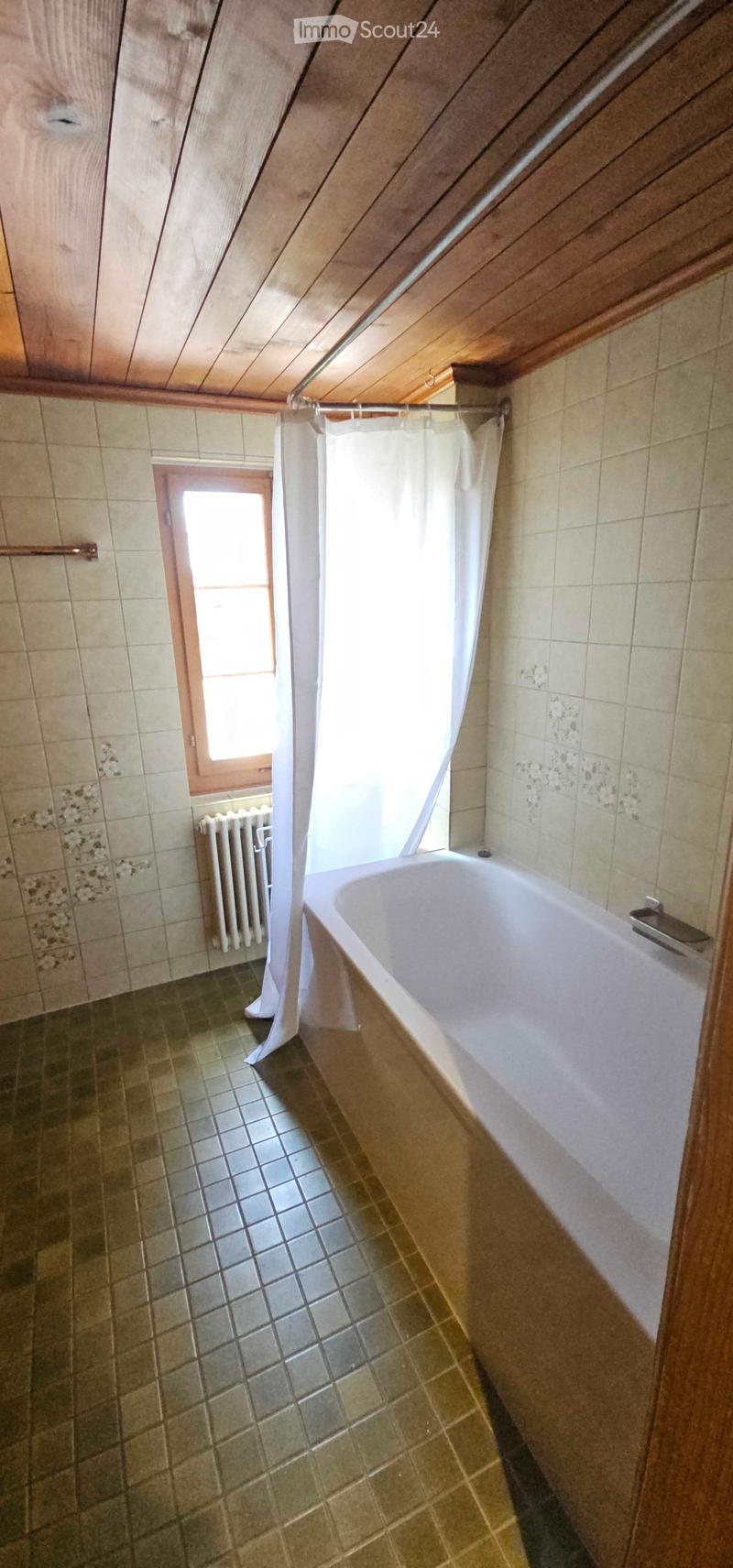 Bathroom with a bathtub, tiled walls, a window, and a radiator. The space features wooden slanted ceilings and a shower curtain.