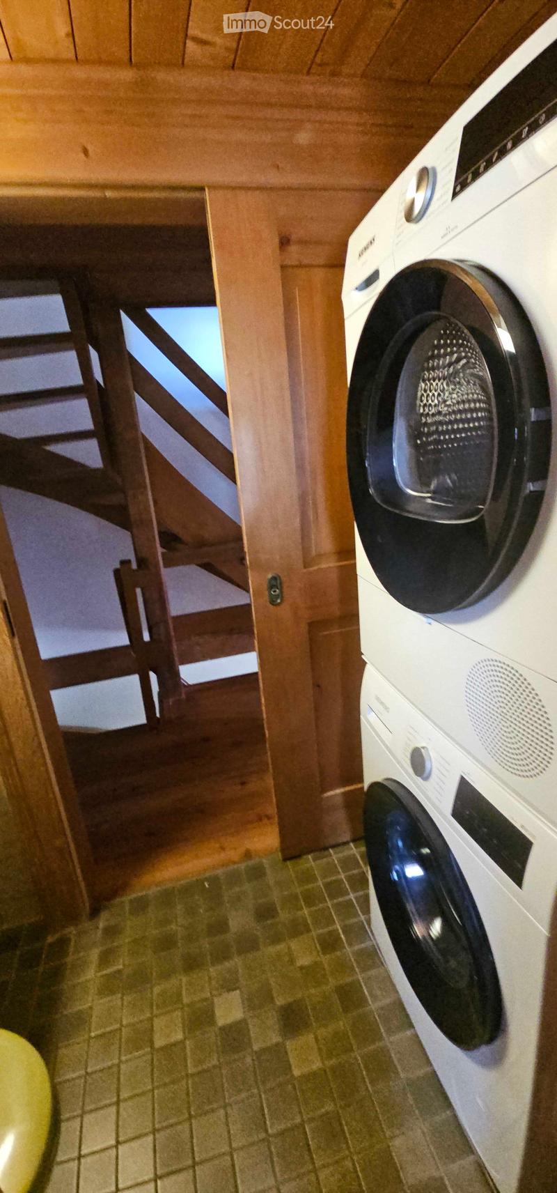 A small room with a washing machine, wooden staircase leading upwards, and tiled floor.