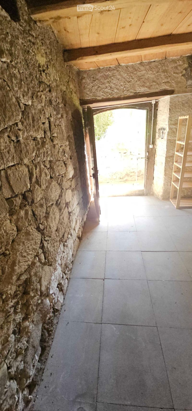 A narrow stone-walled corridor with a wooden door leading to an outdoor area, concrete tiled floor, and exposed wooden beams on the ceiling.
