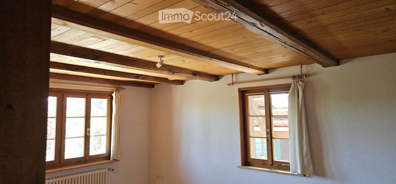 Empty room with wooden ceiling beams, white walls, two windows with wooden frames and curtains, and a radiator under one window.