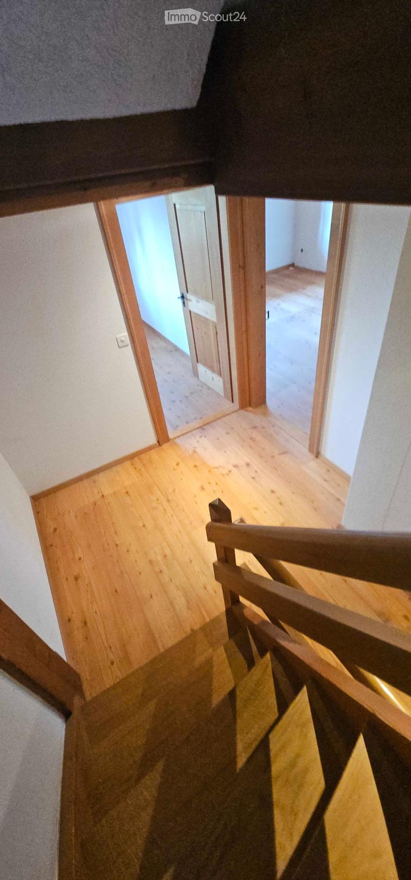 Wooden staircase with a wooden railing, leading to an open doorway with light coming through. The walls are painted white, and the floor is wooden.