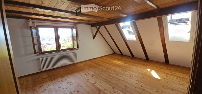 Empty attic room with wooden floor, exposed wooden beams, large windows, and radiator.