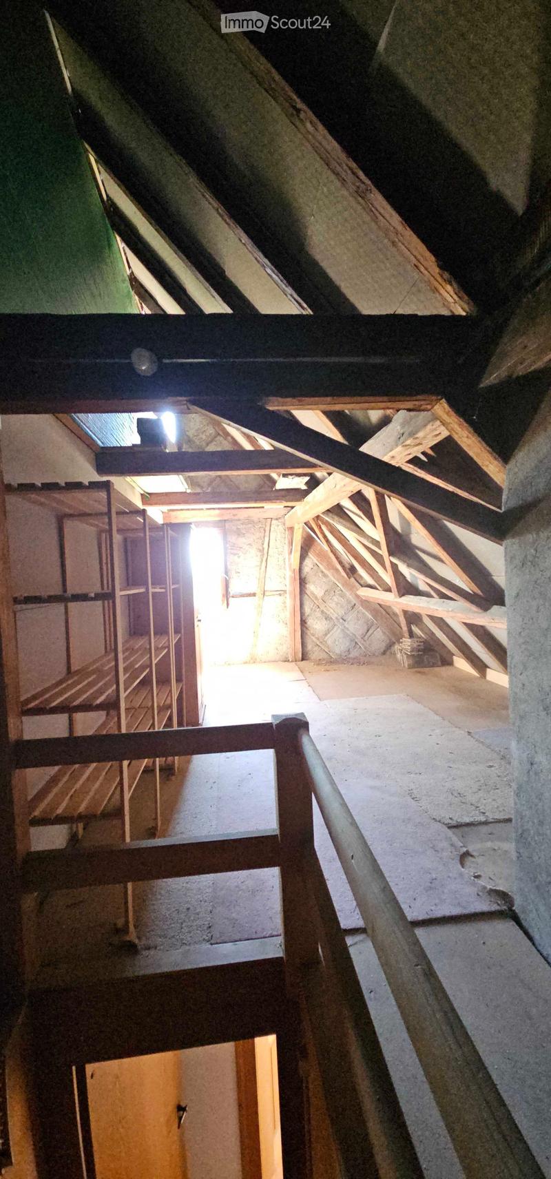 An attic space with exposed wooden beams, concrete walls, and a small skylight allowing natural light to enter. The floor is unfinished concrete, and there is a wooden staircase leading down.