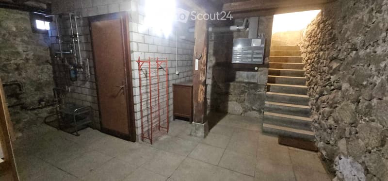 A narrow, dimly lit basement corridor with stone walls, a metal staircase leading upwards, a wooden door, and utility equipment visible.