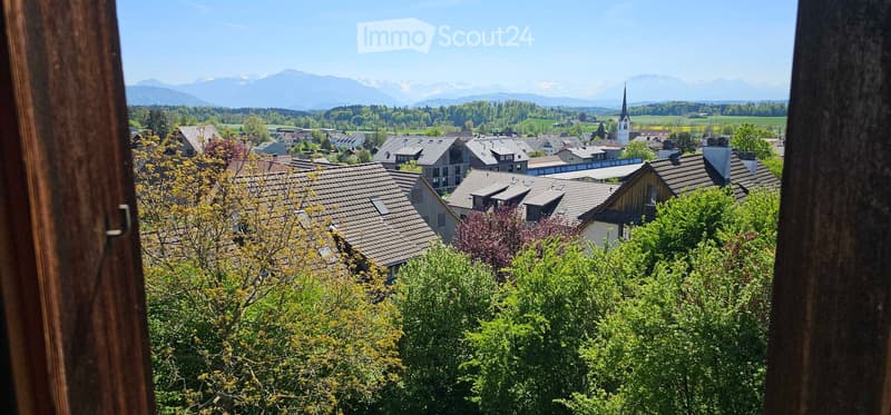 Panoramic view of a village with traditional houses, green trees, and distant mountains under a clear blue sky.