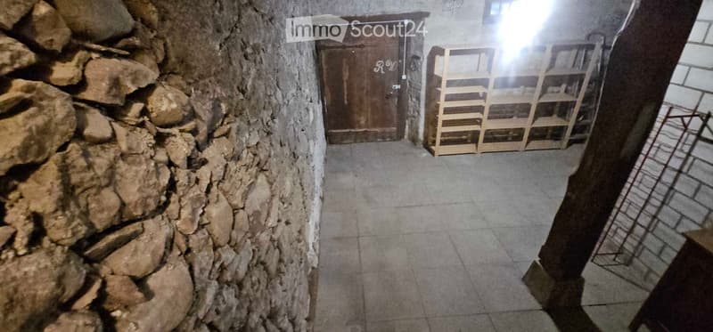 A narrow stone-walled corridor with a wooden door at the end and a wooden shelf unit on the right side.