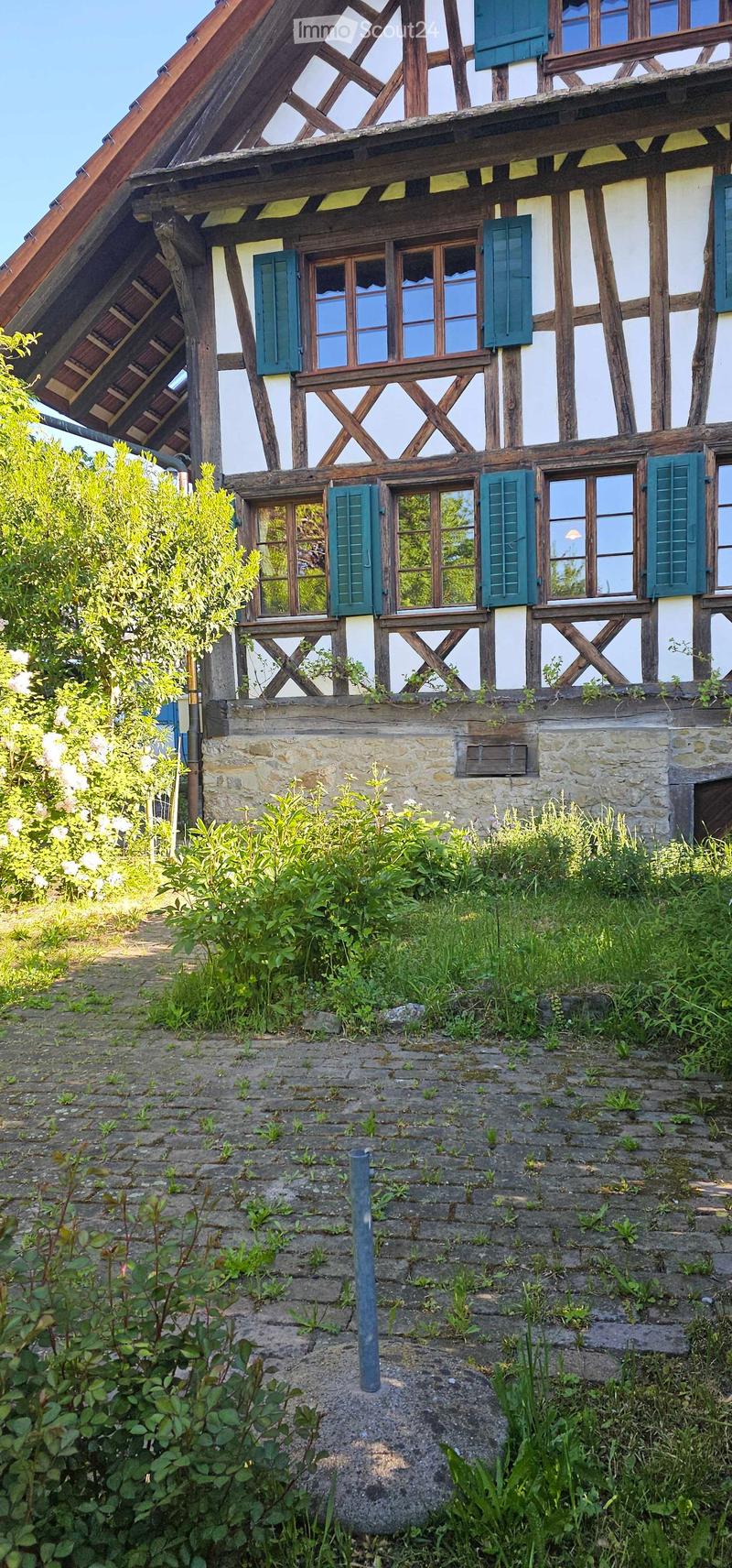 Traditional half-timbered house with white plaster walls, green shutters, and a stone base. The roof features exposed wooden beams and a dormer window. The surrounding area includes overgrown grass and a stone-paved pathway.