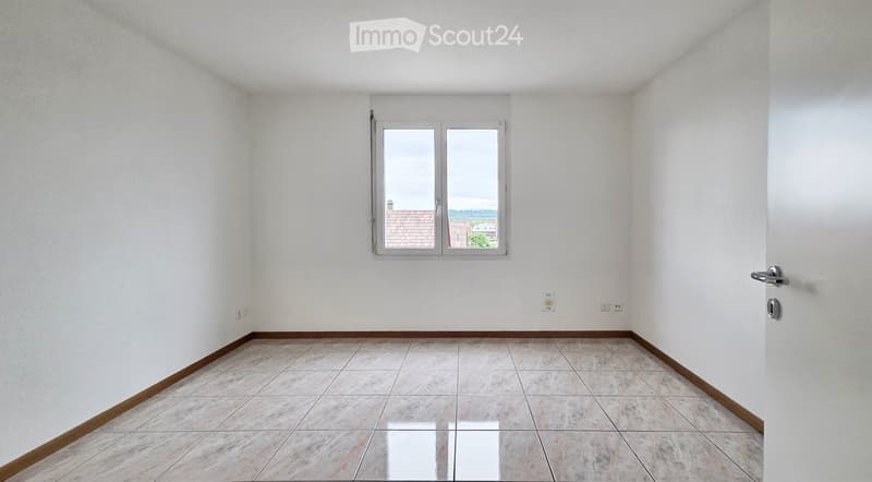 Empty room with white walls, tiled floor, and a window with a view of rooftops and landscape.