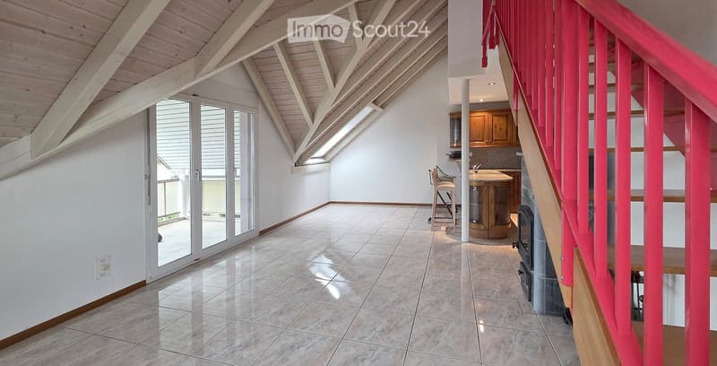 Bright, open room with a high, slanted wooden ceiling, large windows, and a staircase with red railings. Includes a small stool, a kitchen island, and cabinets.